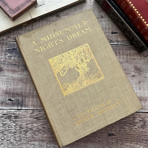 Pode incluir: Uma cópia vintage de "A Midsummer Night's Dream" de William Shakespeare, ilustrada por Arthur Rackham. O livro tem uma capa bege com letras douradas e uma ilustração dourada de uma árvore. Outros livros são visíveis ao fundo.