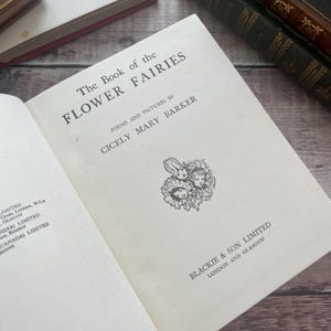 May include: An open book, "The Book of the Flower Fairies" by Cicely Mary Barker, with black text and a fairy illustration. The pages are off-white. Other books are visible in the background on a wooden surface.