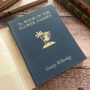 May include: A vintage book titled "The Book of the Flower Fairies" by Cicely M. Barker. The dark blue cover features gold lettering and a gold illustration of a fairy. The book is open, revealing off-white pages. Other books are in the background.