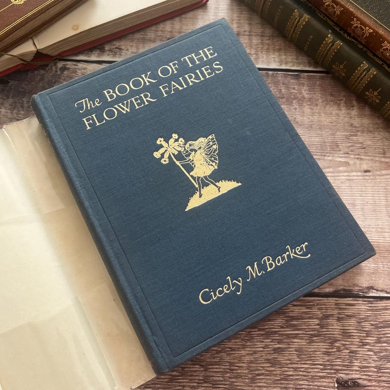 May include: A vintage book titled "The Book of the Flower Fairies" by Cicely M. Barker. The dark blue cover features gold lettering and a gold illustration of a fairy. The book is open, revealing off-white pages. Other books are in the background.