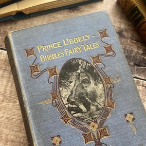 May include: Antique book titled "Prince Ubbely - Bubbles Fairy Tales" with a light blue cover. The cover features gold lettering and an illustration within an ornate frame. Other books are visible in the background.