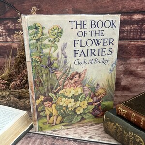 May include: A vintage copy of "The Book of the Flower Fairies" by Cicely M. Barker. The cover shows illustrations of fairies and flowers. The book's title is in blue text. The cover has signs of wear.