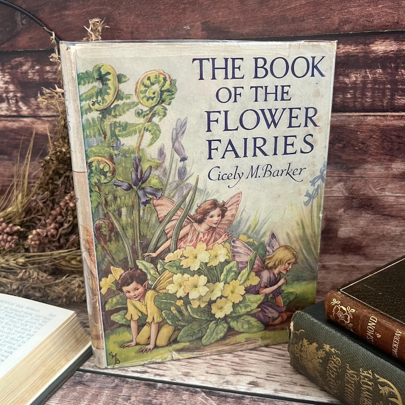 May include: A vintage copy of "The Book of the Flower Fairies" by Cicely M. Barker. The cover shows illustrations of fairies and flowers. The book's title is in blue text. The cover has signs of wear.
