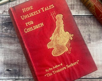 May include: A red hardback book titled Nine Unlikely Tales for Children with gold embossed lettering and a gold illustration of a figure in a long cloak. The cover includes the text By the Author of The Treasure Seekers in gold font.