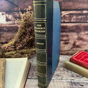 Könnte beinhalten: Ein altes Buch mit dem Titel "The Pilgrim's Progress" von Bunyan. Der Buchrücken ist dunkelgrün mit goldener Schrift. Der blaue Einband und die abgenutzten Kanten weisen auf das Alter des Buches hin.