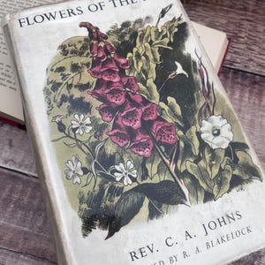 May include: A hardcover book titled Flowers of the Field by Rev. C. A. Johns, edited by R. A. Blakelock. The off-white cover features a detailed botanical illustration of a tall purple foxglove surrounded by smaller white flowers and green foliage.
