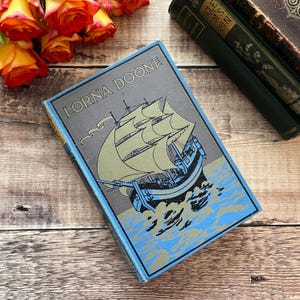 May include: A vintage hardcover book titled "Lorna Doone" with a blue cover and a detailed illustration of a ship with sails on the ocean.