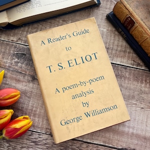 May include: A Reader's Guide to T.S. Eliot book with a light tan cover and teal text. The book is lying on a wooden surface with other books and flowers. The book's title is visible.