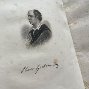 May include: A black and white engraving of a man holding a book. The engraving is on a page with the signature "Oliver Goldsmith" written in black ink.