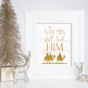 May include: A white framed print with gold lettering that reads "Wise Men still seek HIM". The print features a silhouette of three wise men riding camels, with a star above them. The print is displayed on a white surface with a gold glitter Christmas tree and two silver reindeer figurines.