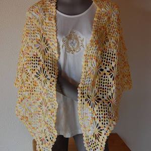 Peut inclure: Un châle blanc en crochet avec des accents jaunes et orange. Le châle est drapé sur une chemise blanche sur un mannequin.