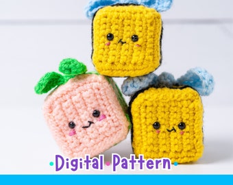 May include: Three crocheted plush toys shaped like cubes with cute faces. The cubes are stacked on top of each other. The top cube is yellow with a blue bow, the middle cube is pink with a green leaf, and the bottom cube is yellow with a blue bow.