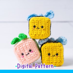 Pode incluir: Três brinquedos de pelúcia de crochê em forma de cubos com rostos fofinhos. Os cubos estão empilhados um em cima do outro. O cubo superior é amarelo com um laço azul, o cubo do meio é rosa com uma folha verde e o cubo inferior é amarelo com um laço azul.