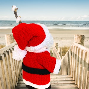 Peut inclure: Un poupon de No&euml;l en crochet portant un bonnet rouge et blanc et une tenue rouge et blanche avec une ceinture noire. Le poupon est debout sur une passerelle en bois surplombant une plage.