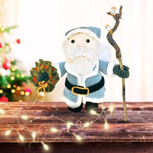 Peut inclure: Un petit P&egrave;re No&euml;l en crochet tenant une couronne verte et un b&acirc;ton en bois avec de petits oiseaux blancs. Le P&egrave;re No&euml;l porte une tenue en crochet bleu et blanc avec une ceinture noire. Il est debout sur une table en bois avec des guirlandes lumineuses blanches.