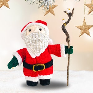 Peut inclure: Un petit P&egrave;re No&euml;l en crochet portant un costume rouge avec des bordures blanches et une barbe blanche. Il tient un b&acirc;ton sur lequel sont perch&eacute;es deux petites oiseaux blancs. La poup&eacute;e est debout sur une surface blanche avec une &eacute;toile dor&eacute;e en arri&egrave;re-plan.