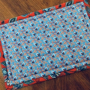 May include: A rectangular quilted placemat with a blue floral pattern center and a red paisley border. The center features a repeating pattern of small blue flowers and orange leaves on a light blue background. The border is a vibrant red with a paisley design.
