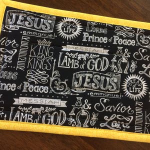 May include: A rectangular black fabric placemat with white script and illustrations, including the words "Jesus," "Lamb of God," and "Prince of Peace." The placemat has a yellow border.