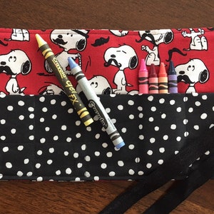 May include: A red and black fabric roll-up case with a black polka dot pattern and a Snoopy print. The case has a black ribbon tie and holds crayons.