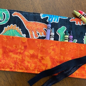May include: A black fabric crayon roll with a dinosaur print and orange lining. The roll has six slots for crayons and a black ribbon tie. The roll is filled with crayons.