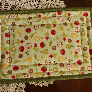 May include: A green quilted placemat with a border of green and yellow fabric. The placemat features a repeating pattern of red apples, yellow pears, and green leaves on a light green background.