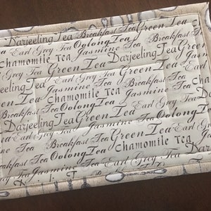 May include: A beige fabric tea coaster with a quilted design. The coaster is decorated with a repeating pattern of various tea types written in black script, including Darjeeling, Earl Grey, Green, Oolong, Jasmine, Chamomile, and Breakfast.