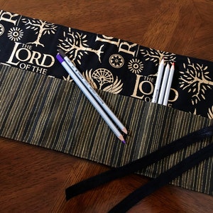 May include: A black and gold pencil roll with a repeating pattern of trees and the words "The Lord of the Rings". The roll is open and has several pencils inside.