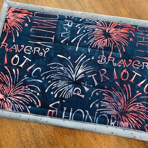 May include: A dark blue fabric placemat with a white and red fireworks design. The placemat has a gray border with white stars. The words "BRAVERY", "PATRIOTIC", and "HONOR" are printed on the fabric.