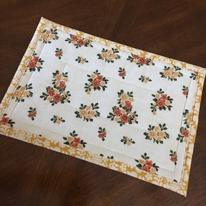 May include: A white quilted placemat with a yellow border and a floral pattern of orange and yellow flowers.