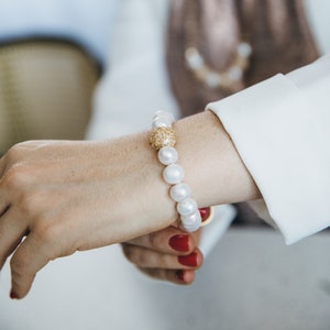 May include: A white pearl bracelet with a gold and crystal accent. The bracelet is on a person's wrist.