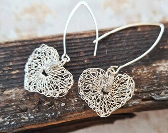 May include: A pair of silver heart-shaped earrings. The earrings feature a delicate, openwork design, with a wire-like texture. The earrings hang from a curved, silver hook. The background is a piece of wood.