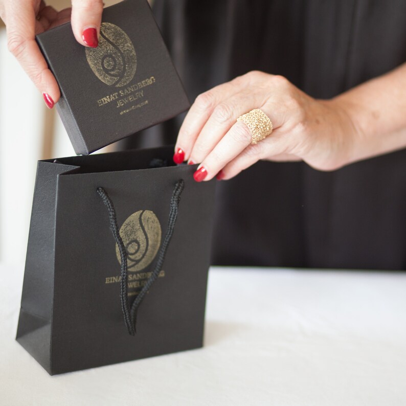 Puede incluir: Una mujer sostiene una caja y una bolsa de regalo negras, ambas con un logotipo dorado y el texto "SINAT SANDBERG JEWELRY". Un anillo dorado adorna su dedo. Las u&ntilde;as est&aacute;n pintadas de rojo.