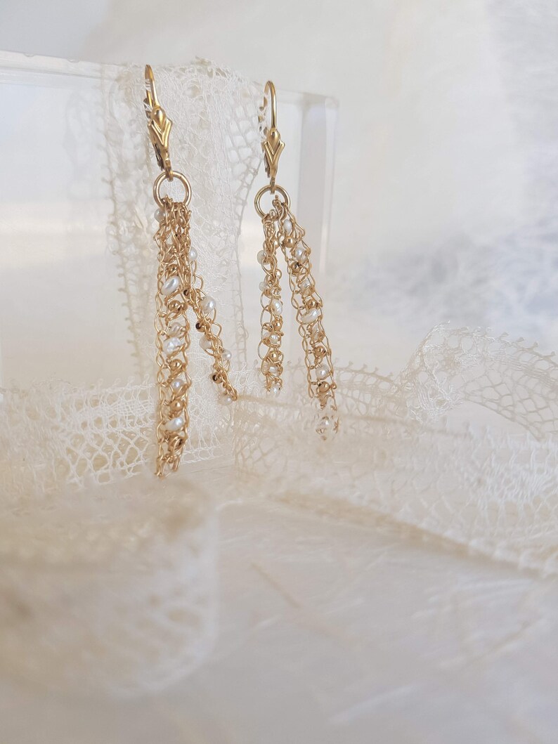 Puede incluir: Pendientes colgantes dorados con delicadas cadenas y peque&ntilde;as perlas blancas. Los pendientes presentan un cierre de palanca y un dise&ntilde;o en cascada, creando un aspecto elegante y con gracia. Los pendientes est&aacute;n sobre un fondo suave y neutro.