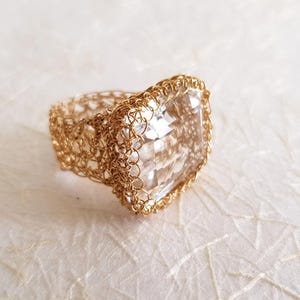 May include: A ring featuring a large, square, clear faceted gemstone set within a band crafted from intricate, woven gold-toned wire. The metal wire creates a delicate, open-work mesh pattern that surrounds the central stone on all sides.