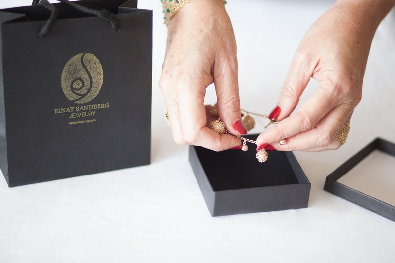 Puede incluir: Un collar dorado con elementos decorativos redondos y texturizados se coloca en una caja de joyer&iacute;a negra. El collar tiene colgantes esf&eacute;ricos y peque&ntilde;os colgantes. Una bolsa de compras negra con un logotipo dorado y el texto "EINAT SANDBERG JEWELRY" est&aacute; en el fondo.