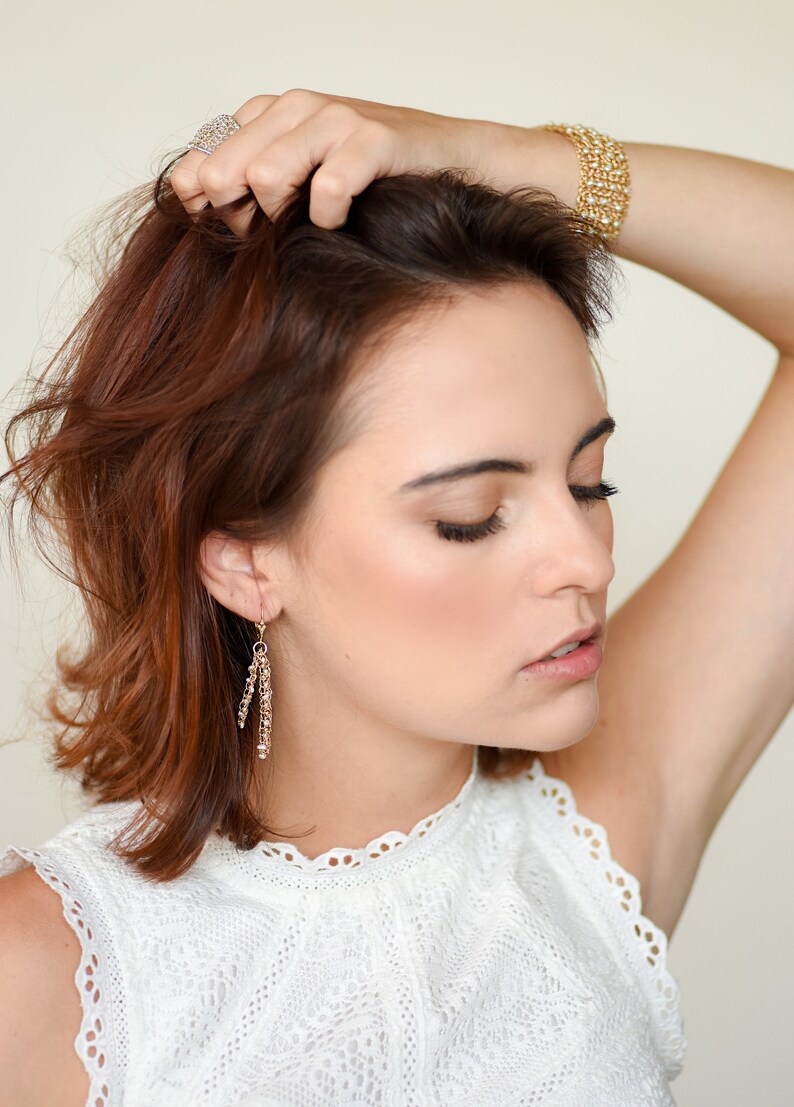 Puede incluir: Una mujer con cabello casta&ntilde;o rojizo lleva pendientes dorados, una pulsera dorada y un anillo plateado. Los pendientes son largos y colgantes. La pulsera tiene un dise&ntilde;o tejido. El anillo es un grupo de peque&ntilde;as piedras.