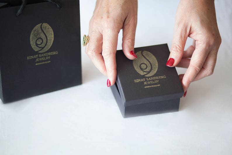 Puede incluir: Una caja de regalo negra y una bolsa de compras a juego con un logotipo dorado y el texto "EINAT SANDBERG JEWELRY". La caja est&aacute; siendo abierta por una persona con esmalte de u&ntilde;as rojo.