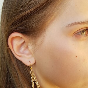 Puede incluir: Pendientes colgantes dorados con m&uacute;ltiples hilos de cadenas delicadas y peque&ntilde;as cuentas blancas. Los pendientes tienen un cierre de gancho y se muestran sobre la piel y el cabello de una persona.