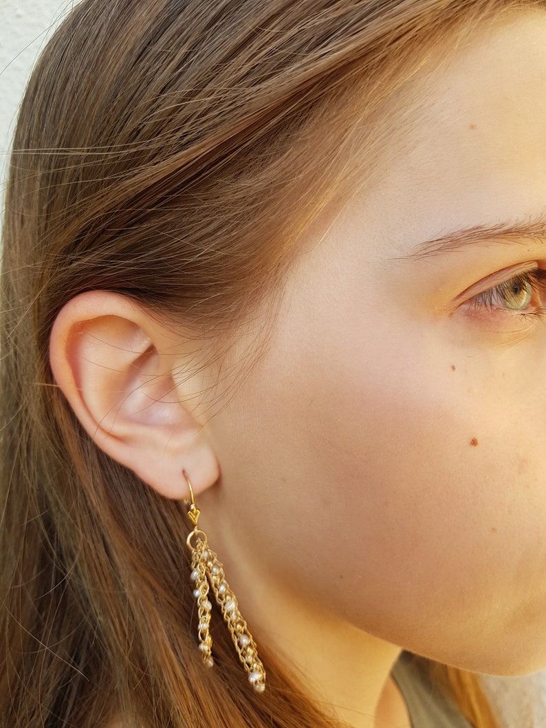 Puede incluir: Pendientes colgantes dorados con m&uacute;ltiples hilos de cadenas delicadas y peque&ntilde;as cuentas blancas. Los pendientes tienen un cierre de gancho y se muestran sobre la piel y el cabello de una persona.