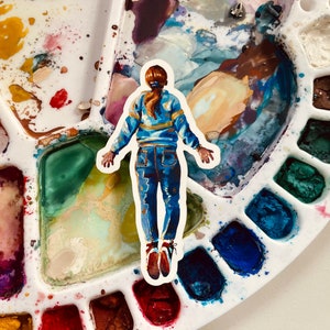 May include: A watercolor painting of a person wearing a blue sweater and jeans, standing on a white paint palette with various colors of paint.