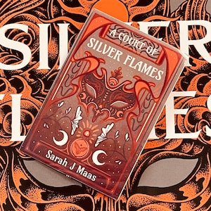 May include: A book cover with a dark brown background and a silver mask with a crown. The title "A Court of Silver Flames" is written in white text. The author's name, "Sarah J Maas", is written in white text at the bottom of the cover.