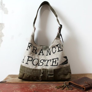 Puede incluir: Un bolso de hombro de lona beige con las palabras "FRANCE POSTE" impresas en negro. El bolso tiene una correa y base verdes, con una cartera de cuero marrón y llaves sobre una superficie de madera.