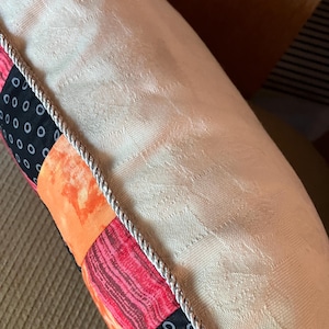 May include: A white pillow with a decorative trim and a patchwork design featuring black, orange, and red fabric.