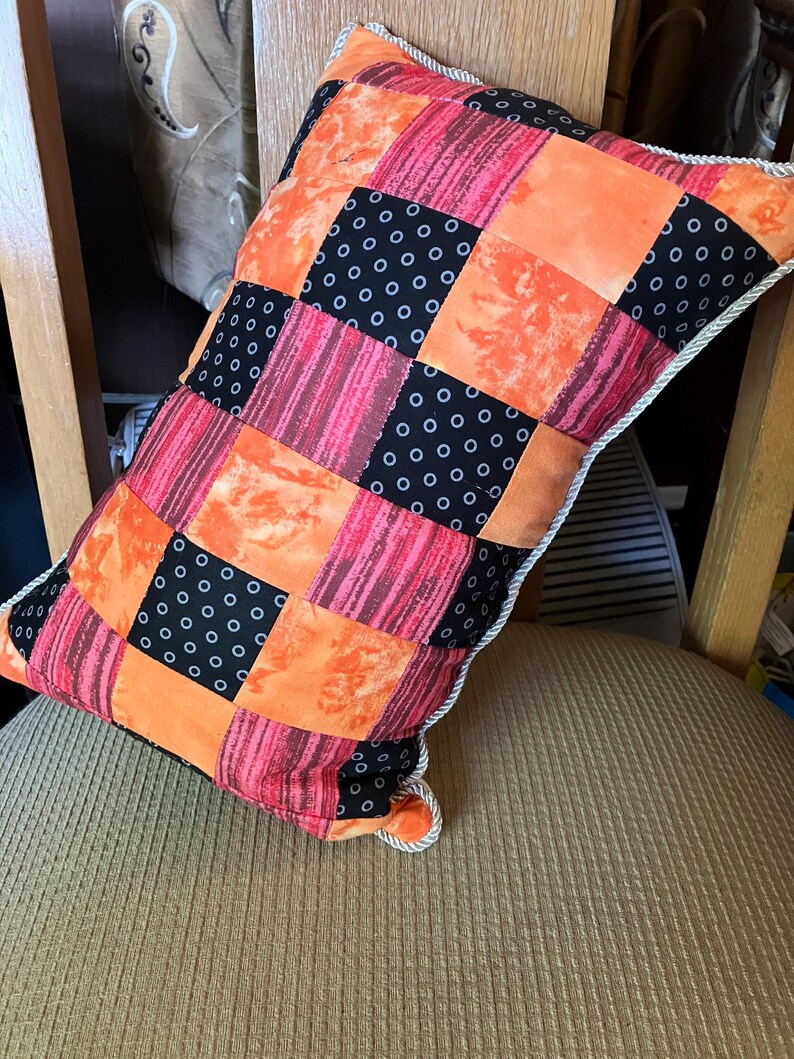 May include: A patchwork pillow with orange, black, and pink fabric squares. The pillow has a white piping around the edges.