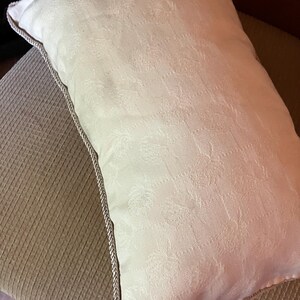 May include: A white pillow with a subtle embossed pattern and a light brown trim. The pillow is sitting on a brown textured surface.