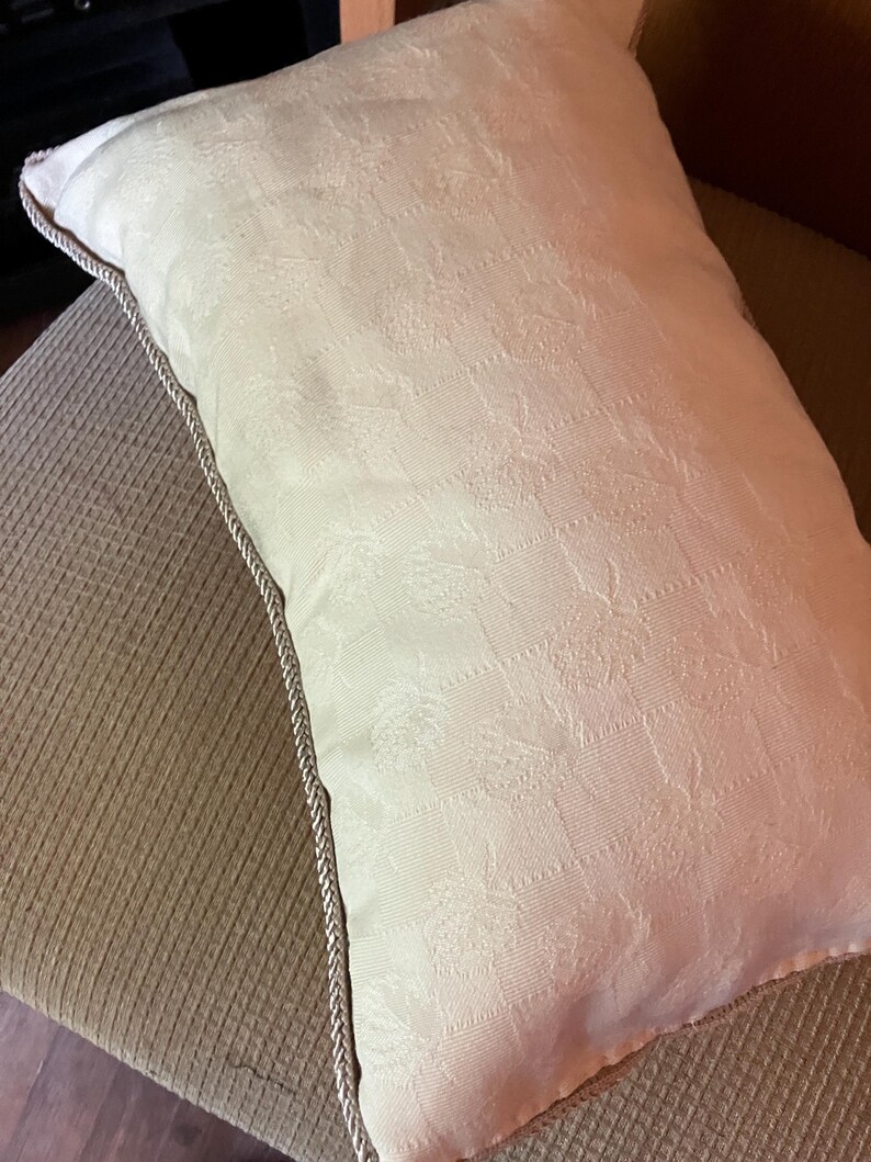 May include: A white pillow with a subtle embossed pattern and a light brown trim. The pillow is sitting on a brown textured surface.