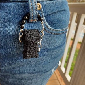 May include: A black crocheted pouch with a silver chain and black beads hanging from a pair of blue jeans.