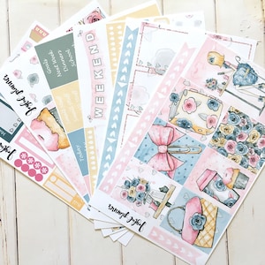 Puede incluir: Una colección de pegatinas para planificador con un diseño de acuarela. Las pegatinas presentan colores pastel rosa, azul y amarillo con motivos florales y de corazón. Algunas pegatinas tienen texto como "Weekend" y "Goals".