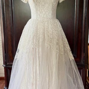 May include: A white floor-length dress featuring a lace bodice with short sleeves and a scoop neckline. The skirt is composed of a satin-like base layer overlaid with a sheer tulle fabric. The lace detailing extends from the bodice onto the skirt.