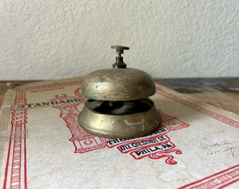 May include: Vintage brass bell with a small handle on top. The bell has a tarnished, aged appearance, with hints of green oxidation. It sits on a vintage sheet music book with red and black text. The bell is a classic decorative item.